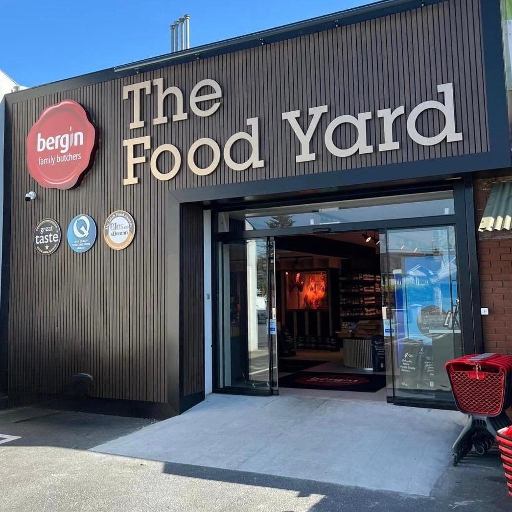food_yard1 The image shows the entrance to a store called The Food Yard, which appears to be operated by Bergin Family Butchers. The exterior has dark vertical paneling with large, bold, cream-colored lettering for the store name. On the left side of the entrance, there are three circular award-style plaques, including a "Great Taste" award and others indicating quality recognition.</p>
<p>The entrance features wide glass sliding doors, revealing part of the store interior with shelves and displays. A red shopping cart is visible to the right of the doorway, suggesting it’s a grocery or specialty food store. The overall appearance is modern and clean.